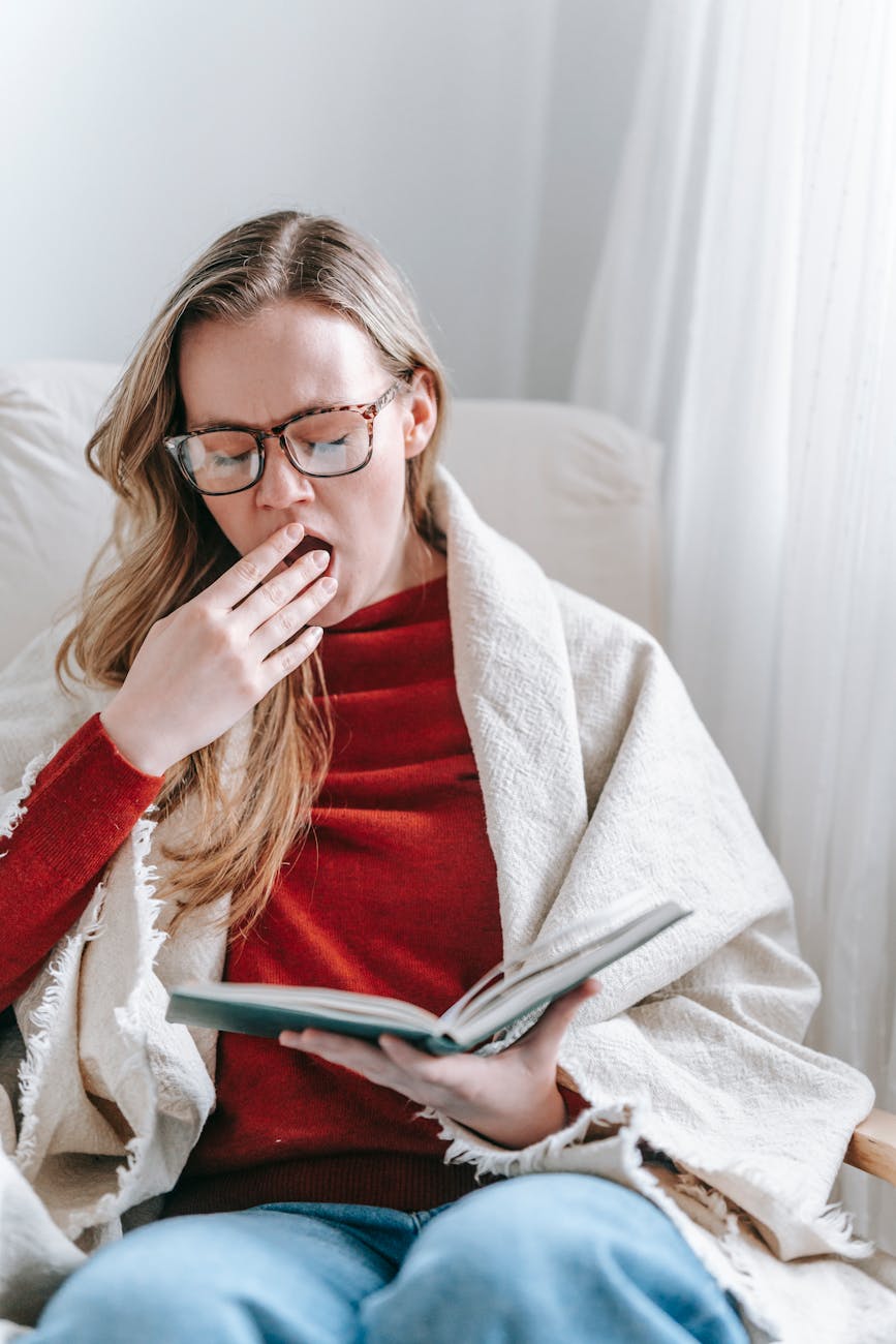 exhausted young woman yawning while reading textbook in armchair